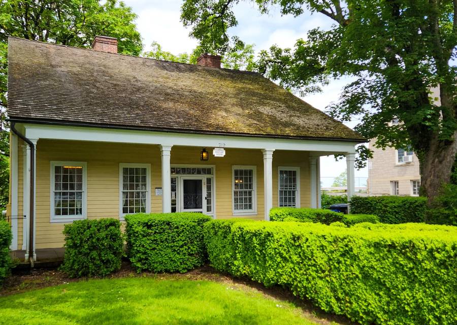 The Barclay House, a yellow single storey cottage with a deep front porch, a mossy shingle roof and a clipped hedge along the lawn.