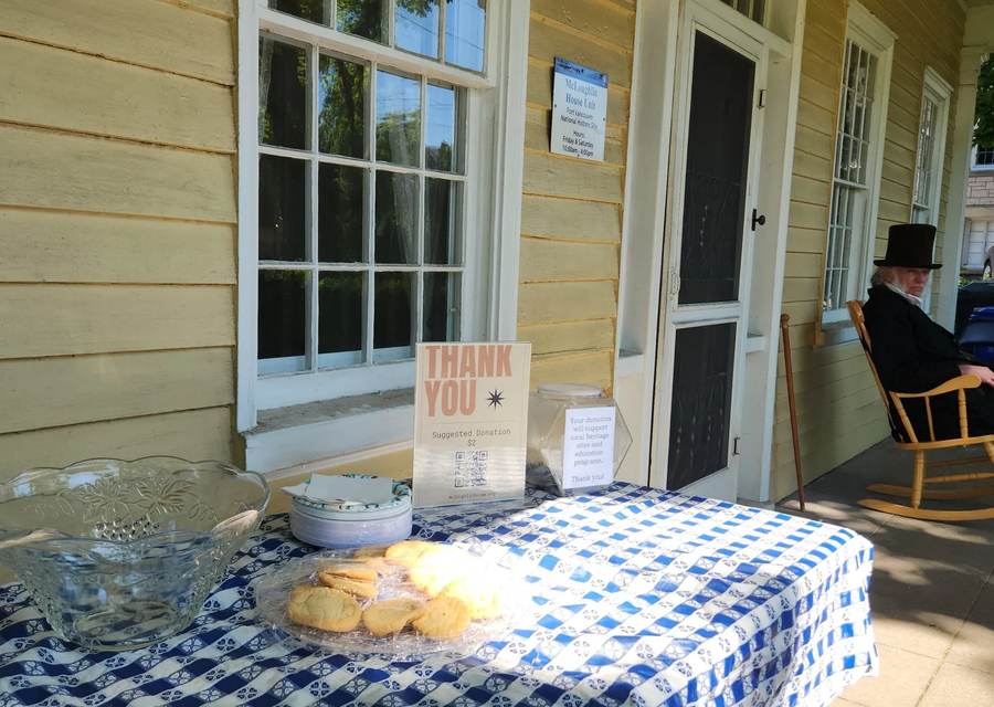 A refreshments table with a punch bowl and cookies set up on the Barclay House porch, with a costumed interpreter in a top hat seated in a rocking chair beside the door.