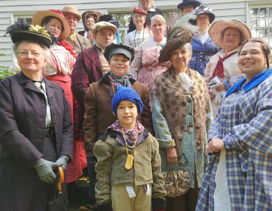 Fifteen volunteers in nineteenth century dress, including two children, gathered on the steps of a white clapboard house.